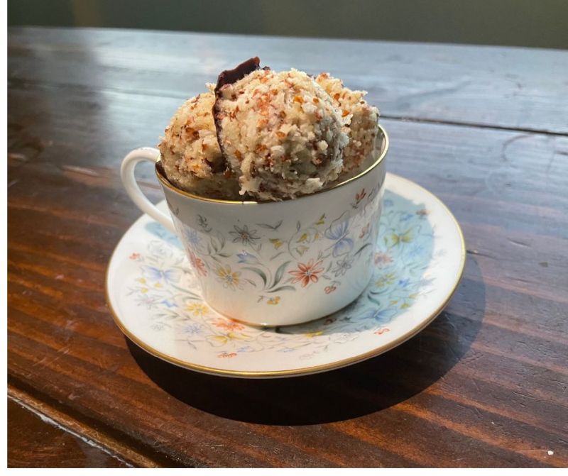 A scoop of ice cream topped with crumbs and a piece of chocolate, served in a floral teacup on a wooden table garnished with asparagus.