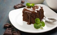 A slice of gluten-free chocolate cake with chocolate sauce and mint leaves on a white plate, accompanied by pieces of chocolate and a milk jug.