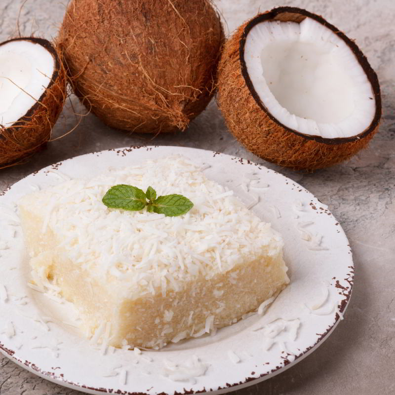 A slice of coconut cake garnished with almondine on a plate, with whole and halved coconuts in the background.
