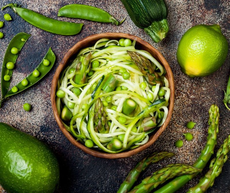A bowl of zucchini noodles garnished with peas and asparagus amidst fresh limes and vegetables on a textured surface, alongside vegan coconut cake.