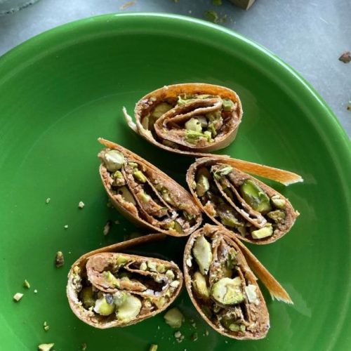 A close-up of five rolled pistachio pastries on a green plate exudes temptation akin to guilt-free gingerbread cookies. Sliced to reveal layers filled with crushed pistachios, they blend golden and green hues with scattered pistachio pieces inviting you to indulge without hesitation.