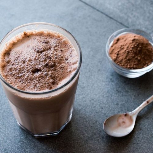 A glass of frothy chocolate milkshake on a gray countertop sits beside a small bowl of cocoa powder, echoing the rich swirls reminiscent of a decadent chocolate marble cheesecake, with a spoon ready for indulgence.