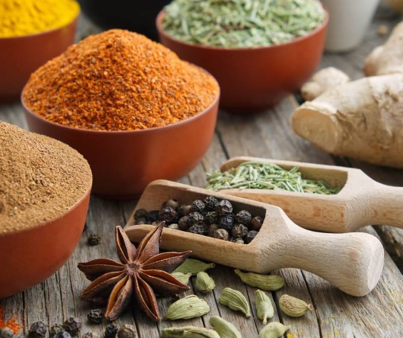 A rustic wooden table displays bowls of various spices alongside a chocolate marble cheesecake. Wooden scoops hold black peppercorns and dried herbs, while star anise, cardamom pods, and ginger root are scattered around the scene, adding a delightful aroma to the cozy setting.