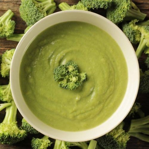A bowl of creamy green broccoli soup garnished with a small piece of broccoli in the center. Fresh broccoli florets are scattered around the bowl on a wooden surface, offering a hint of earthy delight similar to that found in spicy macadamia walnut dip.
