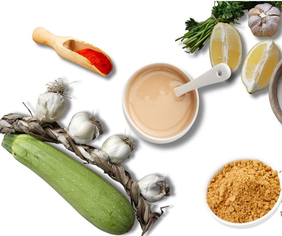 A flat lay of heart-friendly paté ingredients on a white background, featuring zucchini, garlic braid, a bowl of sauce with a spoon, lemon halves, leafy herbs, a wooden scoop with red spice, and brown crumbs.