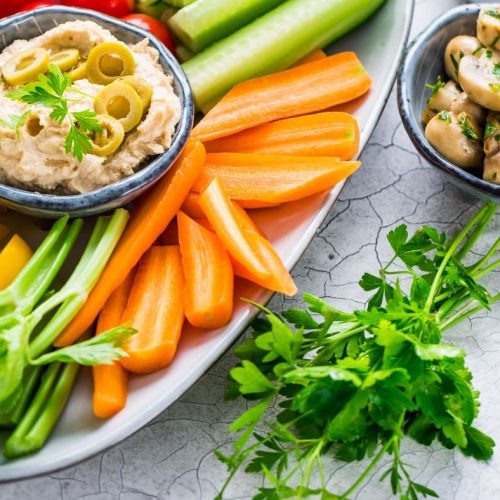 A vibrant vegetable platter with celery, carrots, and yellow peppers accompanies a heart-friendly paté alongside a bowl of hummus topped with olives. A separate dish holds marinated mushrooms, and a sprig of fresh parsley is placed on the side.