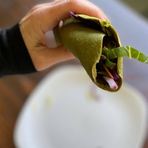 A hand holding a Green Flax Wrap filled with leafy greens and vegetables, above a white plate on a wooden table.
