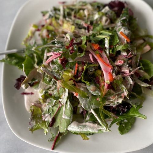 A fresh mixed green salad with sliced red onions, shredded carrots, and leafy greens, dressed in a creamy sauce, accompanies a hemp milk green smoothie. Served on a white rectangular plate, with a fork placed on the side.