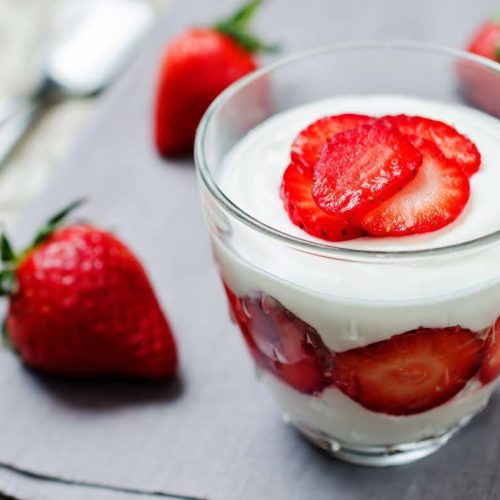 A glass cup filled with layers of yogurt and fresh strawberry slices creates a tempting Strawberry Parfait, placed on a gray cloth with whole strawberries and a spoon in the background.