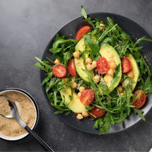 A black plate of salad with arugula, sliced avocado, cherry tomatoes, and chickpeas sits next to a bowl of grated cheese or nutritional yeast—perfect alongside Cheezy Broccoli Bites—on a dark background.
