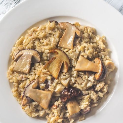A white plate filled with creamy risotto topped with sliced mushrooms and a drizzle of vegan queso, placed on a light wooden surface.