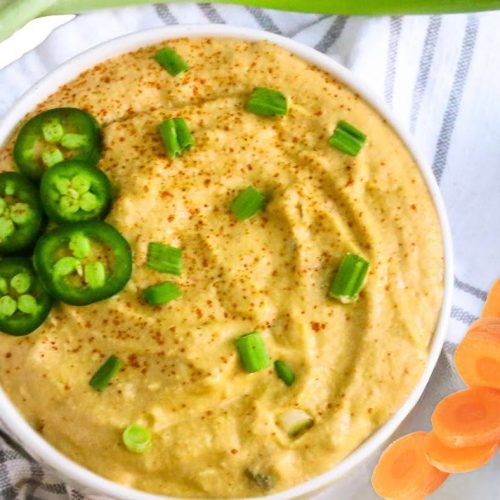 A bowl of creamy dip garnished with sliced jalapeños, chopped green onions, and a sprinkle of seasoning sits on a striped cloth, served alongside carrot slices, green onions, and fresh Vegan Sushi Rolls.