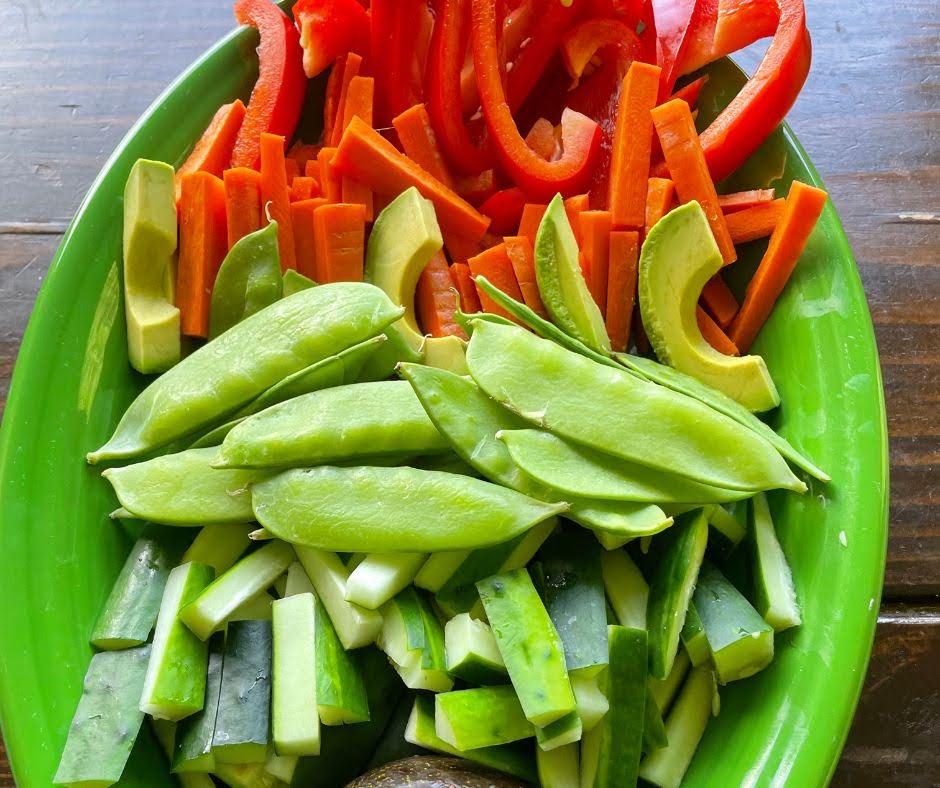 A green platter filled with sliced red bell peppers, carrots, avocado, sugar snap peas, and cucumber sticks arranged in colorful sections—perfect for a Hormone-Happy Salad.