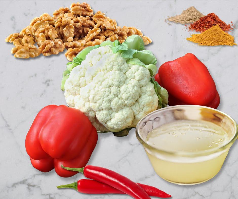 A marble surface displays ingredients for Vegan Sushi Rolls: walnuts, a head of cauliflower, two red bell peppers, two red chili peppers, a bowl of oil, and small piles of ground spices.