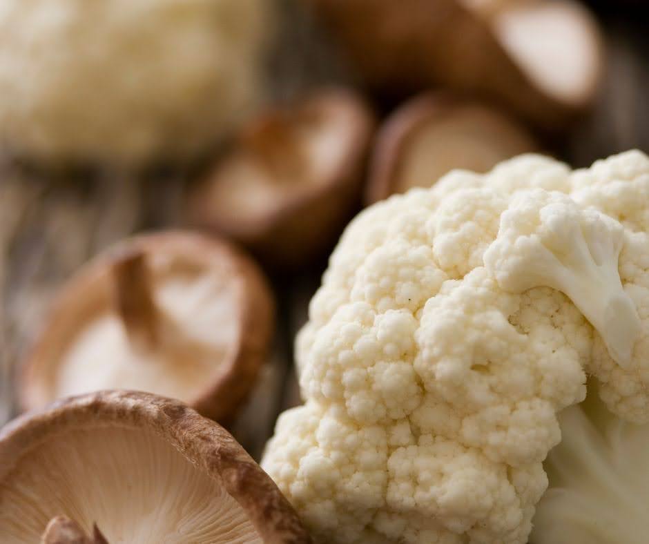 Close-up of a fresh cauliflower floret next to several sliced mushrooms on a rustic surface. Perfect for vegan queso recipes, the cauliflower takes center stage, while the mushrooms are softly blurred in the background.