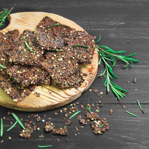 A wooden board on a dark surface holds several flaxseed crackers, garnished with fresh rosemary sprigs and served with Creamy Cauliflower Alfredo Sauce. Some flaxseed crackers and rosemary are scattered around the board.