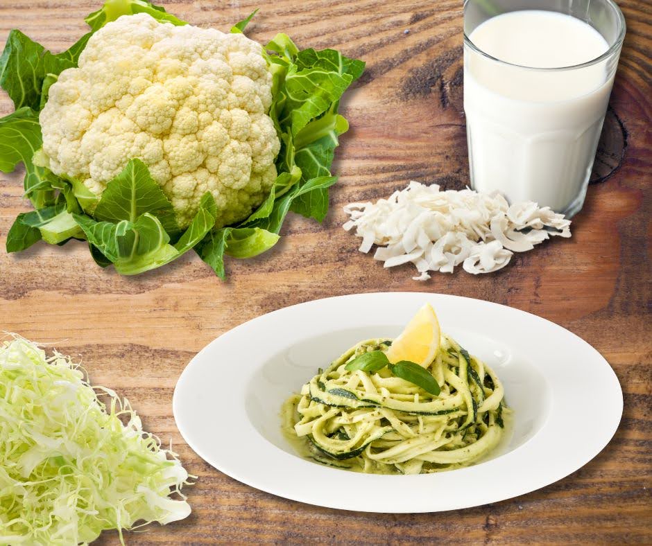 A plate of pasta with pesto and a lemon wedge is on a wooden table, surrounded by a head of cauliflower, Cauliflower Rice with Shiitake Mushrooms & Coconut Sauce, a glass of milk, shredded cheese, and shredded lettuce.