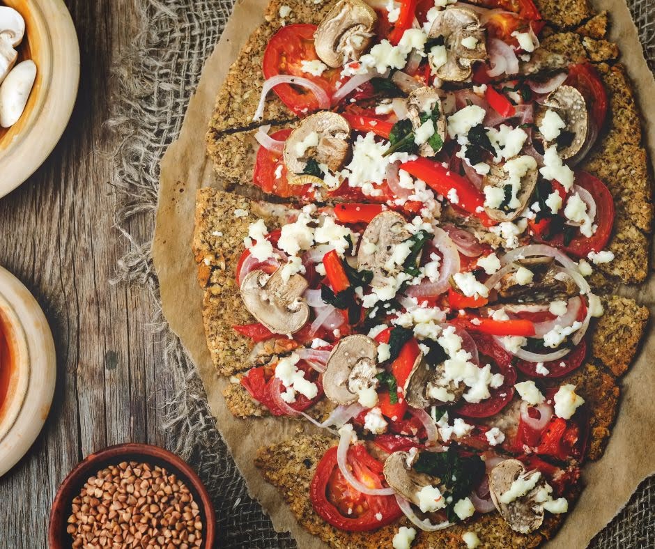 A rustic, homemade vegetable pizza with creamy cauliflower Alfredo sauce sits on parchment paper, topped with tomatoes, mushrooms, onions, red peppers, spinach, and crumbled cheese on a wooden table surrounded by bowls of seeds and mushrooms.