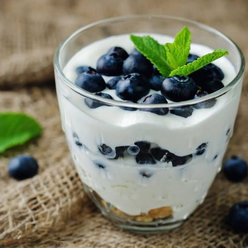 A glass filled with layers of creamy yogurt and fresh blueberries, topped with a sprig of mint—perfect alongside your Adrenal Energy Tea. Some blueberries and a mint leaf are scattered on the burlap fabric background.