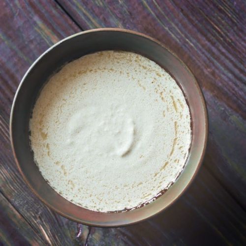 A metal bowl filled with fine, off-white Cauliflower Rice with Shiitake Mushrooms & Coconut Sauce sits on a rustic wooden surface. The smooth powder is slightly mounded in the center, ready to be enjoyed.