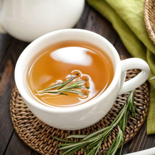 A white cup filled with herbal tea, garnished with a sprig of fresh rosemary, sits on a woven coaster. Nearby on the wooden table are Vegan Chia Crackers, extra rosemary sprigs, and a green napkin.