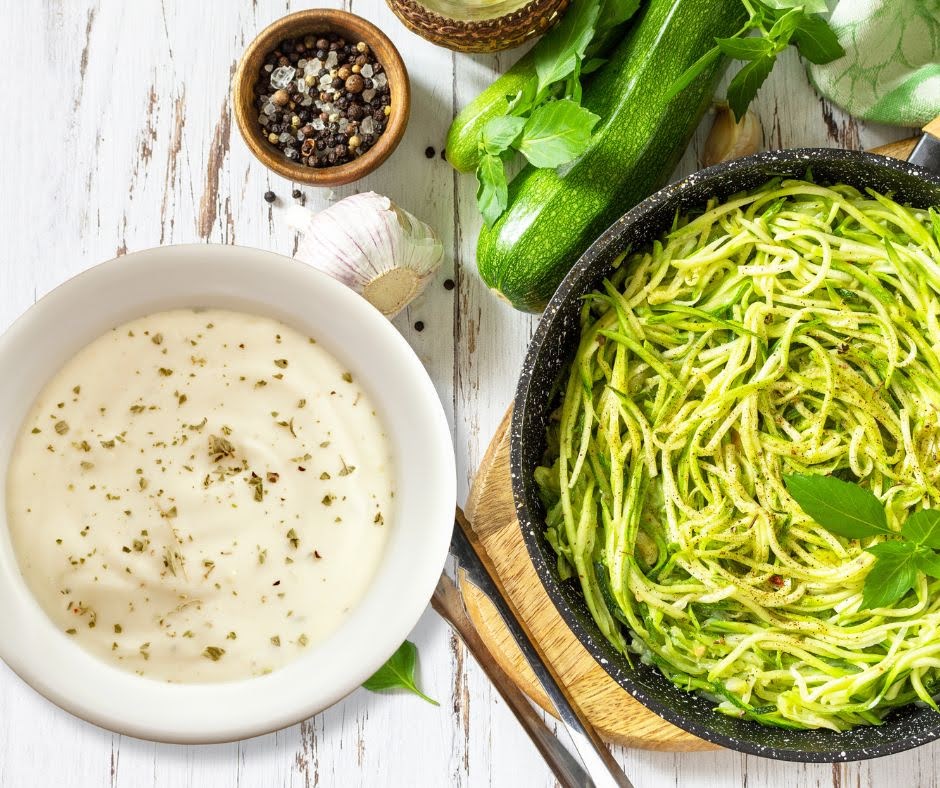 A bowl of creamy white sauce garnished with herbs sits next to a pan of spiralized zucchini noodles and fresh basil on a rustic white wooden table, alongside ingredients for Cauliflower Rice with Shiitake Mushrooms & Coconut Sauce.