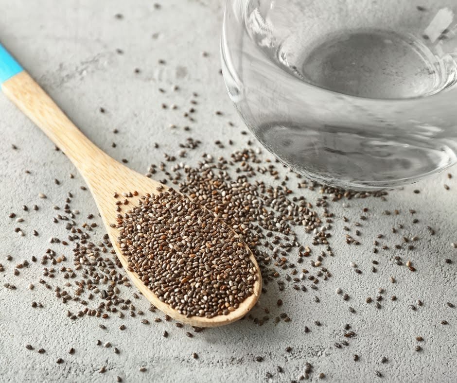 Wooden spoon filled with chia seeds, scattered on a light gray surface next to a clear glass of water—perfect for adding to your Creamy Cauliflower Alfredo Sauce for extra texture and nutrition.