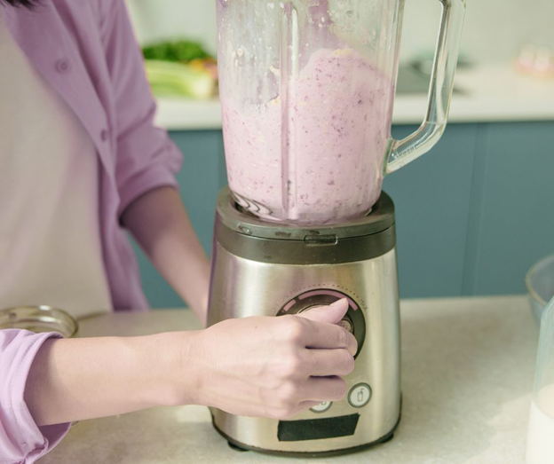 A person in a light purple shirt uses a blender filled with a thick pink mixture, possibly a smoothie, on a kitchen counter with some vegetables in the background.