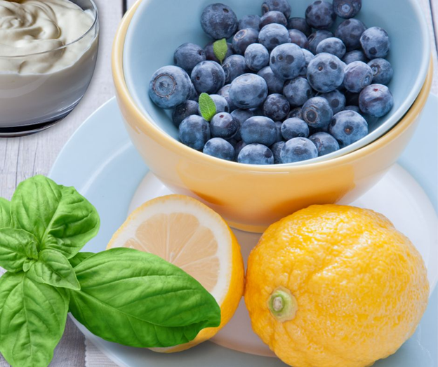 A bowl of fresh blueberries sits atop a plate with two lemons and basil leaves, with a glass of creamy yogurt in the background on a light wooden table.