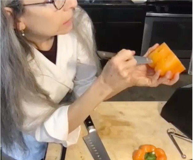 A woman with long gray hair and glasses holds an orange bell pepper and points a knife at it in a kitchen. A cutting board with a bell pepper and knives is in front of her.