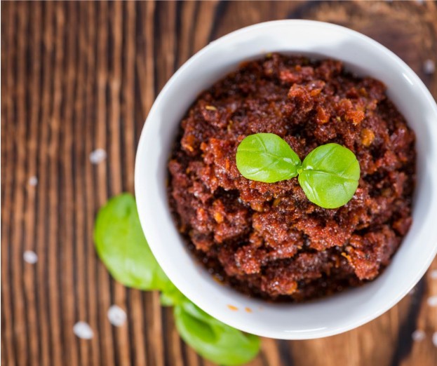 A white bowl filled with red sun-dried tomato pesto, garnished with two fresh basil leaves, sits on a wooden table with scattered salt and basil leaves nearby.