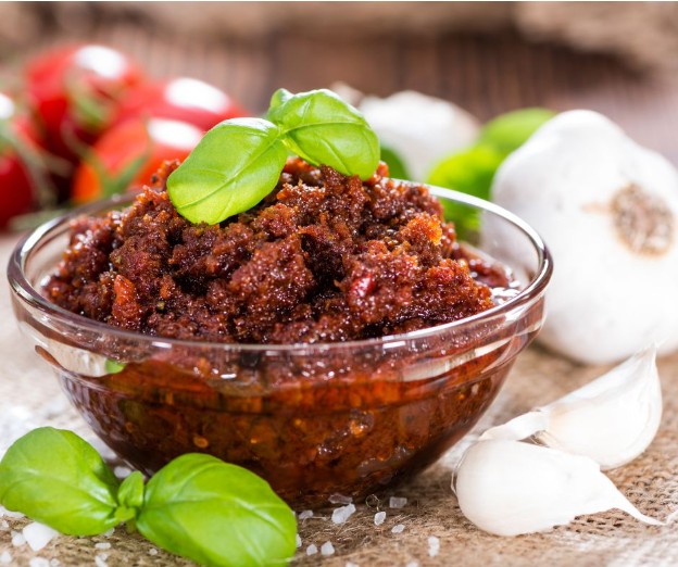 A glass bowl filled with red pesto sauce, garnished with fresh basil leaves. In the background, there are whole tomatoes, a head of garlic, and loose garlic cloves on a rustic surface.