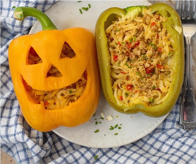 Two bell peppers stuffed with noodles and vegetables are on a plate. One yellow pepper is carved like a jack-o’-lantern, and the other green pepper is halved. Plate rests on a checkered cloth.