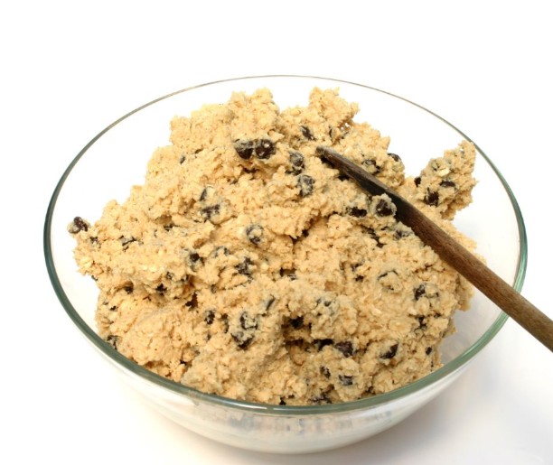 A clear glass bowl filled with cookie dough mixed with raisins, with a wooden spoon resting inside, set against a white background.