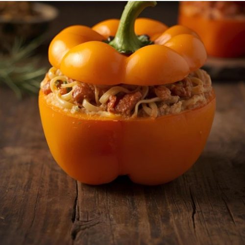 A bright orange bell pepper with its top cut off, stuffed with spaghetti and a tomato-based meat sauce, sits on a rustic wooden surface.