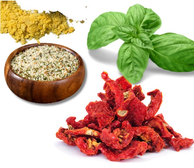 A pile of yellow nutritional yeast, fresh green basil leaves, a wooden bowl of mixed seasoning, and a heap of sun-dried tomatoes on a white background.