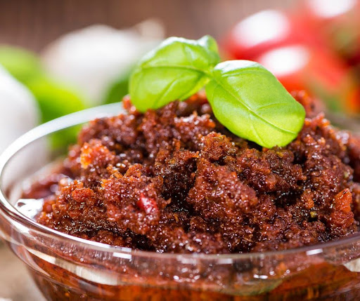 A close-up of sun-dried tomato pesto in a glass bowl, garnished with fresh basil leaves. The texture is chunky and rich, with visible pieces of sun-dried tomato and herbs.