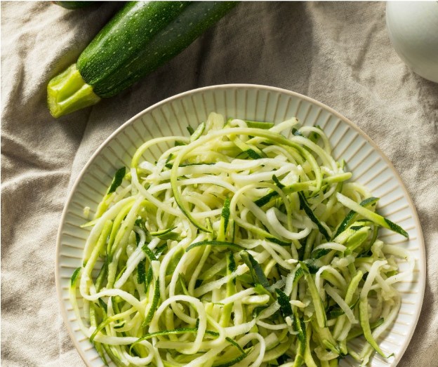 A plate of raw zucchini noodles, also known as zoodles, sits on a light-colored cloth with two whole zucchinis next to it.