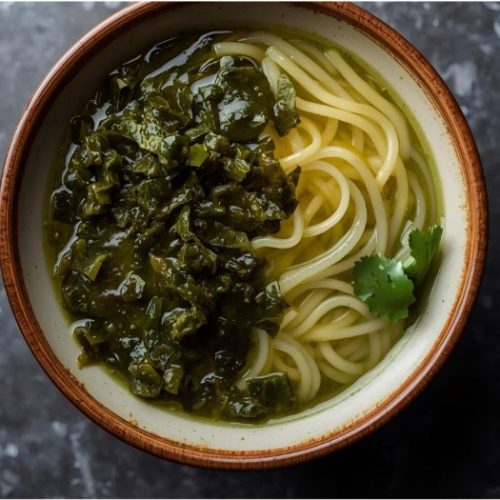 A bowl of noodles topped with a green leafy sauce, garnished with a sprig of cilantro, set on a dark marbled surface.