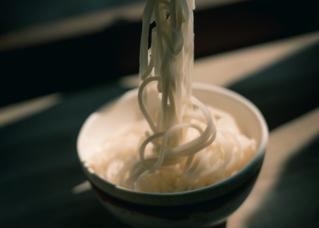 A bowl of white noodles with some noodles being lifted out, set in soft natural light with a blurred background.