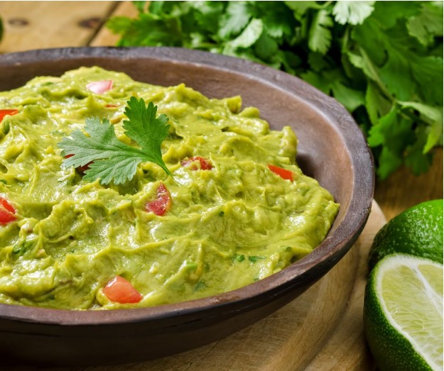 A wooden bowl filled with creamy guacamole, garnished with a cilantro leaf and pieces of tomato. Fresh cilantro and a halved lime are placed beside the bowl on a wooden surface.