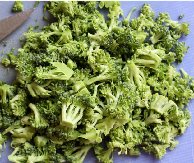 Chopped broccoli florets scattered on a light blue cutting board, with part of a knife visible in the upper left corner.