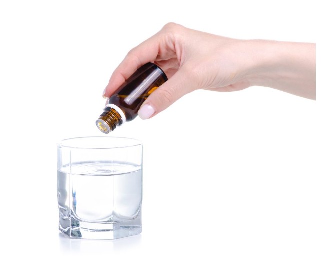 A hand holds a small brown bottle, pouring drops of liquid into a clear glass of water against a white background.