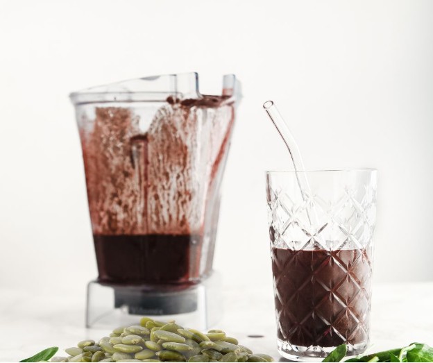 A blender jar containing a thick, dark purple smoothie sits beside a tall glass filled with the same smoothie. The glass has a glass straw, and green pumpkin seeds are scattered in the foreground.