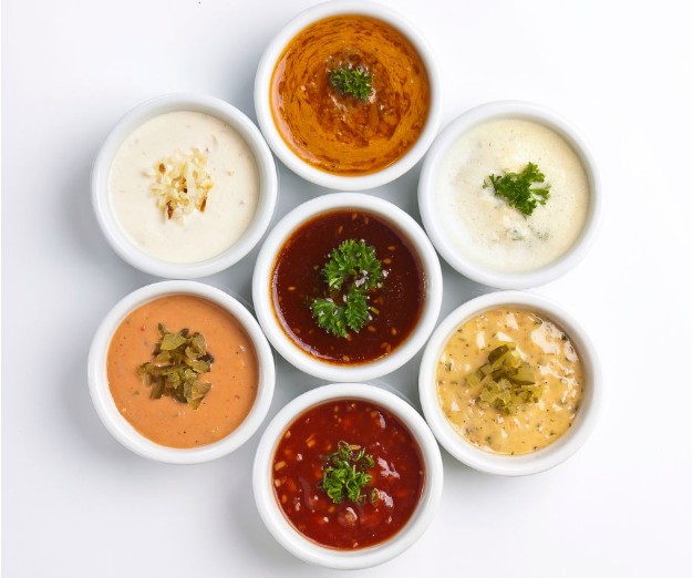 Seven small white bowls arranged in a circle, each filled with a different dipping sauce. Most are topped with a garnish of herbs or chopped vegetables, displayed on a white background.