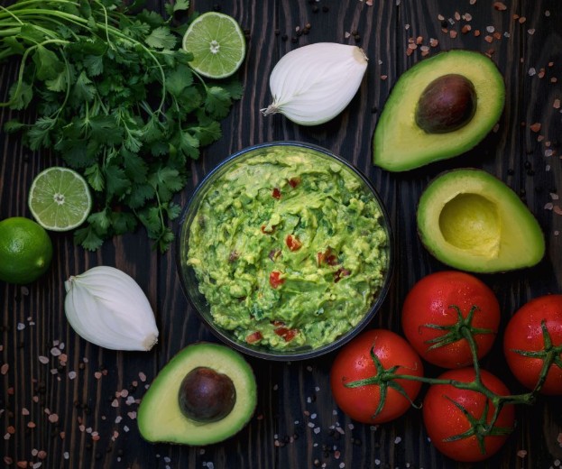 A bowl of guacamole surrounded by fresh ingredients: avocados, tomatoes on the vine, onions, lime halves, and cilantro, all arranged on a dark wooden surface.