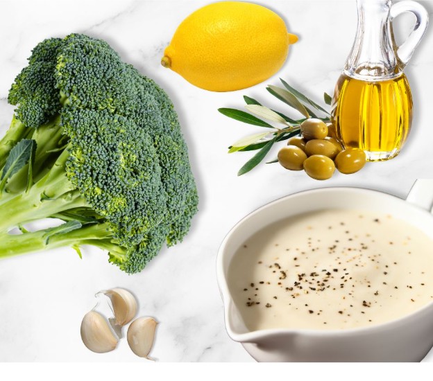 Broccoli, a lemon, a bottle of olive oil with olives and leaves, a white sauce in a bowl topped with black pepper, and garlic cloves, all arranged on a white marble surface.