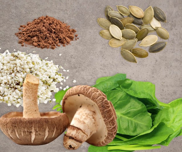 A cluster of fresh spinach leaves, two mushrooms, a pile of shelled hemp seeds, pumpkin seeds, and a mound of brown powder are arranged on a gray surface.