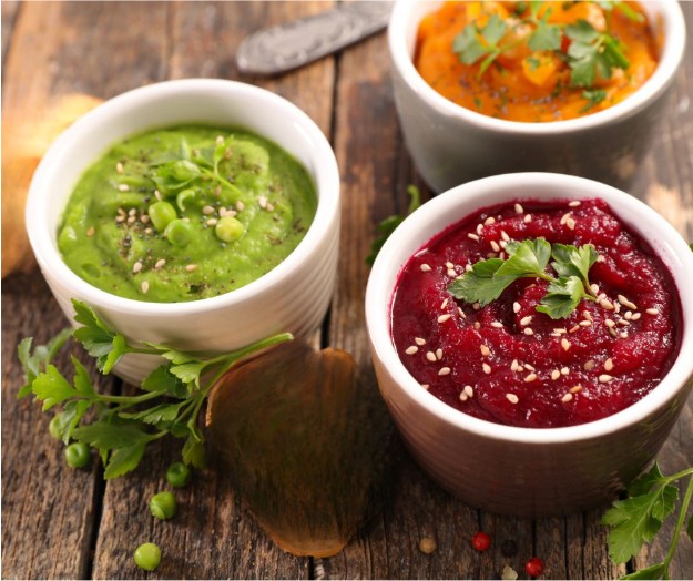 Three small bowls of colorful dips on a wooden table: green dip garnished with peas and herbs, red dip with parsley and sesame seeds, and orange dip topped with herbs, with scattered parsley and peas nearby.
