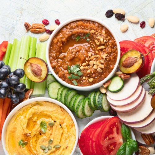 A colorful platter with bowls of hummus and red dip, surrounded by sliced cucumbers, tomatoes, carrots, celery, radishes, grapes, plums, nuts, asparagus, and fresh basil leaves.